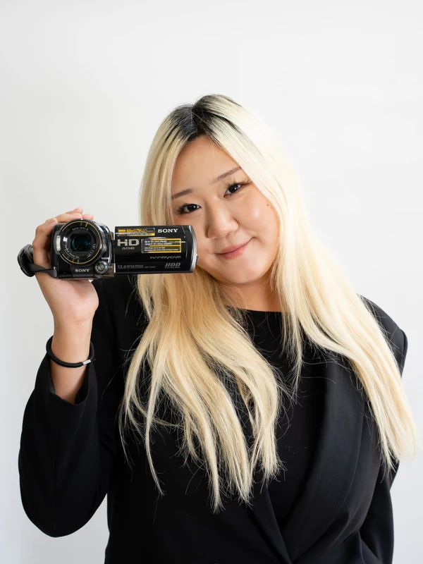 Hannah Chang in a black blazer, holding a Sony HD Handycam camcorder up beside her face against a plain white background.